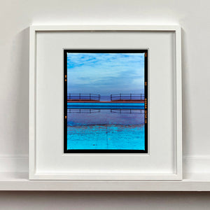 White framed photograph by Richard Heeps. The blue bay at Llandudno, cut across the middle with path and railings with a gap right in the middle.