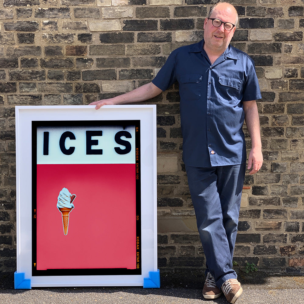 Photograph by Richard Heeps.  Richard Heeps holds a white framed print. At the top of the print, black letters spell out ICES and below is depicted a 99 icecream cone sitting left of centre against a green coloured background.  