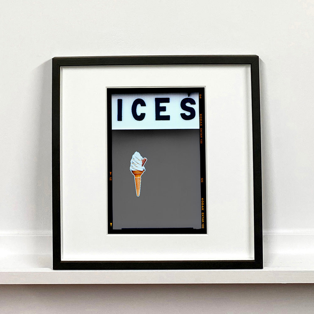 Black framed photograph by Richard Heeps.  At the top black letters spell out ICES and below is depicted a 99 icecream cone sitting left of centre against a grey coloured background.  