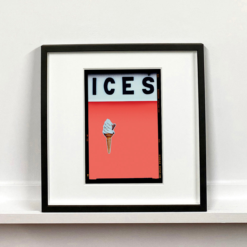 Black framed photograph by Richard Heeps.  At the top black letters spell out ICES and below is depicted a 99 icecream cone sitting left of centre against a melondrama red orange coloured background.  