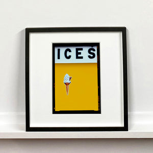 Black framed photograph by Richard Heeps.  At the top black letters spell out ICES and below is depicted a 99 icecream cone sitting left of centre against a mustard yellow coloured background.  