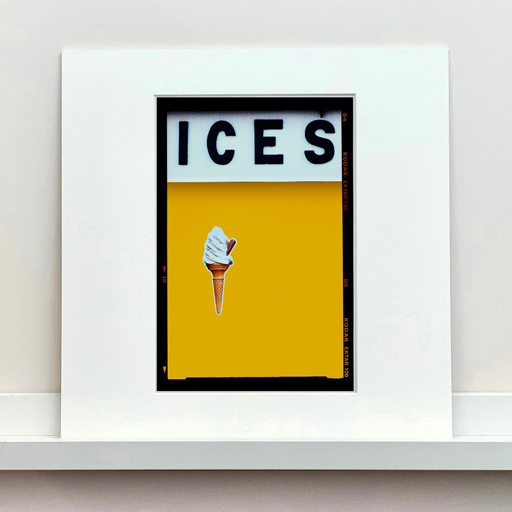 Mounted photograph by Richard Heeps.  At the top black letters spell out ICES and below is depicted a 99 icecream cone sitting left of centre against a mustard yellow coloured background.  