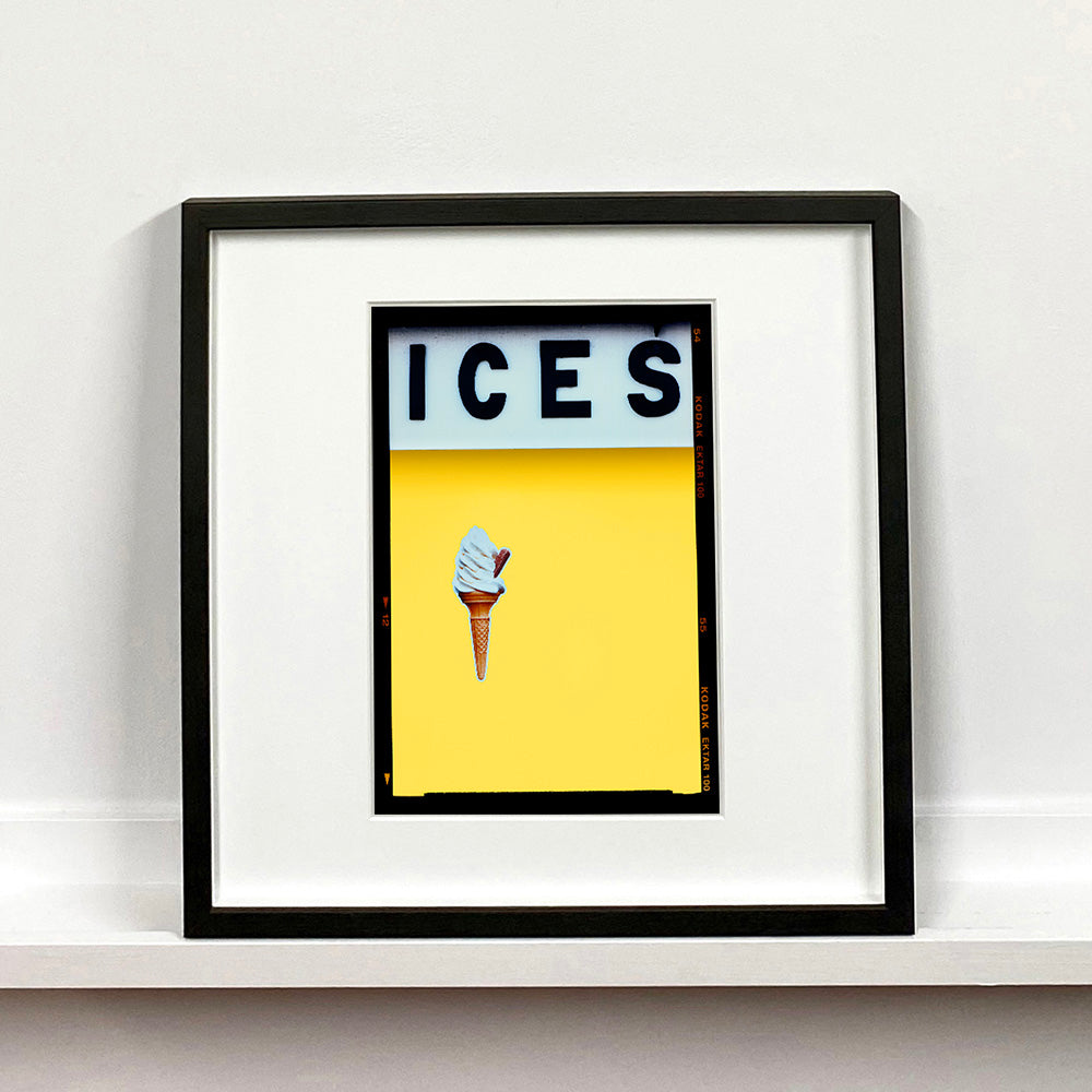 Black framed photograph by Richard Heeps.  At the top black letters spell out ICES and below is depicted a 99 icecream cone sitting left of centre against a sherbet yellow coloured background.  