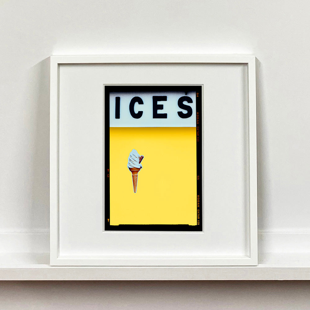 White framed photograph by Richard Heeps.  At the top black letters spell out ICES and below is depicted a 99 icecream cone sitting left of centre against a sherbert yellow coloured background.  