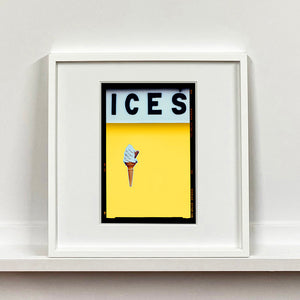 White framed photograph by Richard Heeps.  At the top black letters spell out ICES and below is depicted a 99 icecream cone sitting left of centre against a sherbet yellow coloured background.  