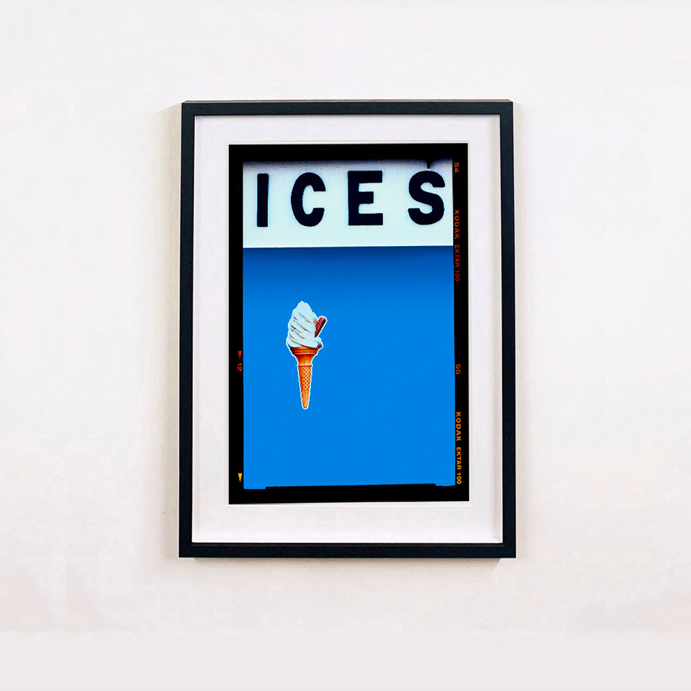 Black framed photograph by Richard Heeps.  At the top black letters spell out ICES and below is depicted a 99 icecream cone sitting left of centre against a sky blue coloured background.  