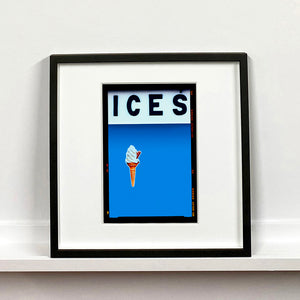 Black framed photograph by Richard Heeps.  At the top black letters spell out ICES and below is depicted a 99 icecream cone sitting left of centre against a sky blue coloured background.  