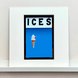 Mounted photograph by Richard Heeps.  At the top black letters spell out ICES and below is depicted a 99 icecream cone sitting left of centre against a sky blue coloured background.  