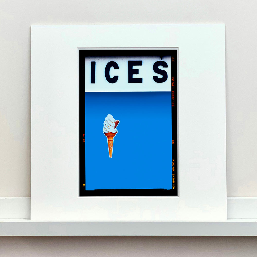 Mounted photograph by Richard Heeps.  At the top black letters spell out ICES and below is depicted a 99 icecream cone sitting left of centre against a sky blue coloured background.  