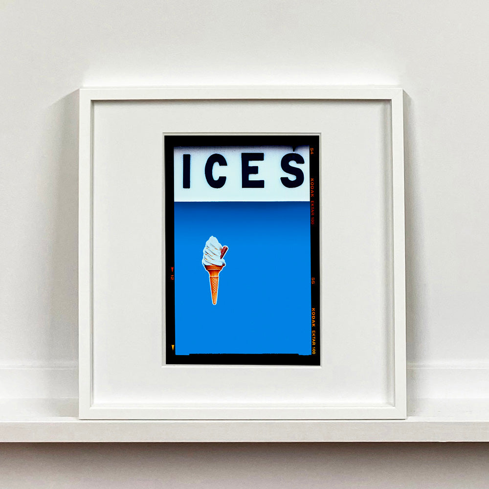 White framed photograph by Richard Heeps.  At the top black letters spell out ICES and below is depicted a 99 icecream cone sitting left of centre against a sky blue coloured background.  