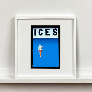White framed photograph by Richard Heeps.  At the top black letters spell out ICES and below is depicted a 99 icecream cone sitting left of centre against a sky blue coloured background.  