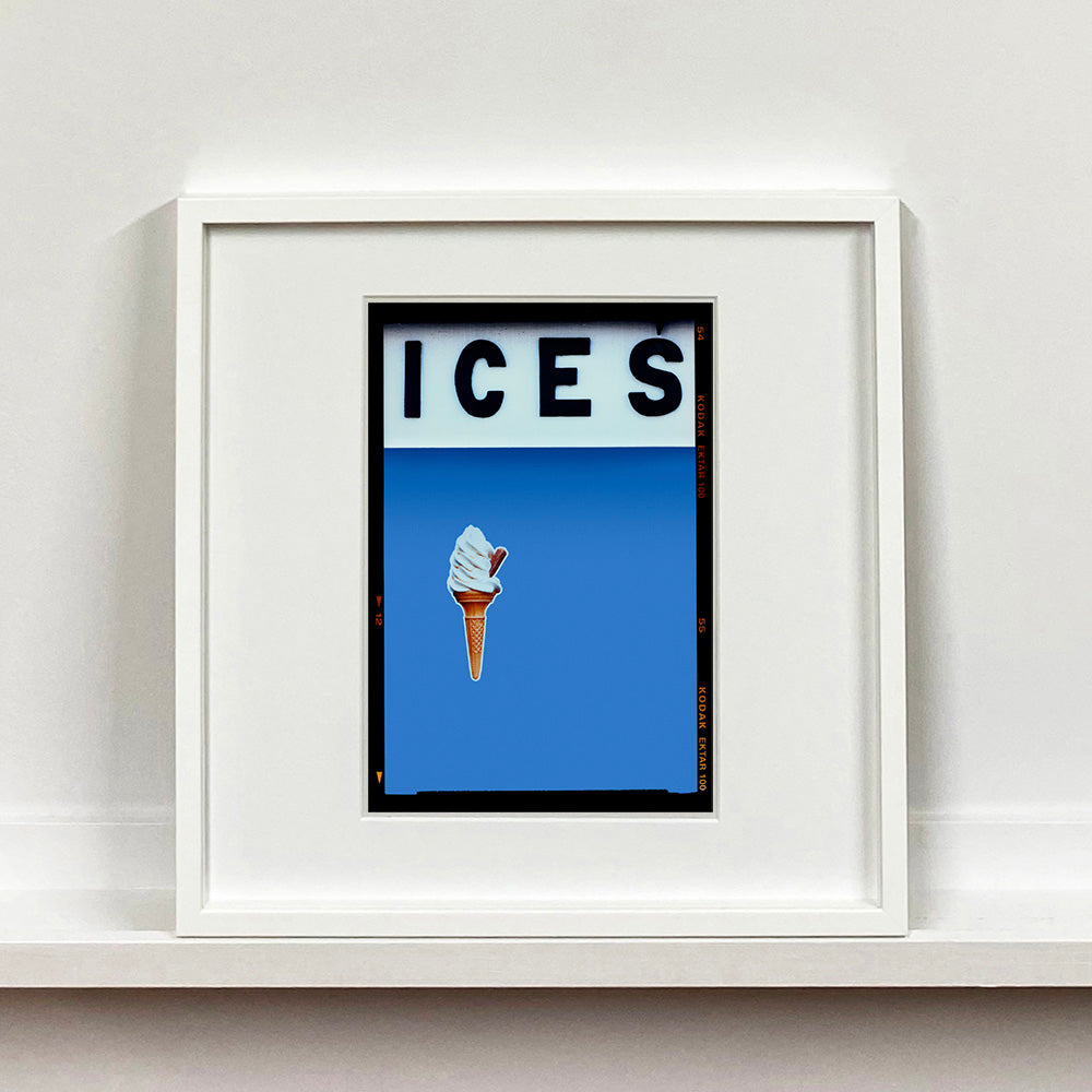 White framed photograph by Richard Heeps.  At the top black letters spell out ICES and below is depicted a 99 icecream cone sitting left of centre against a Baby Blue coloured background.  