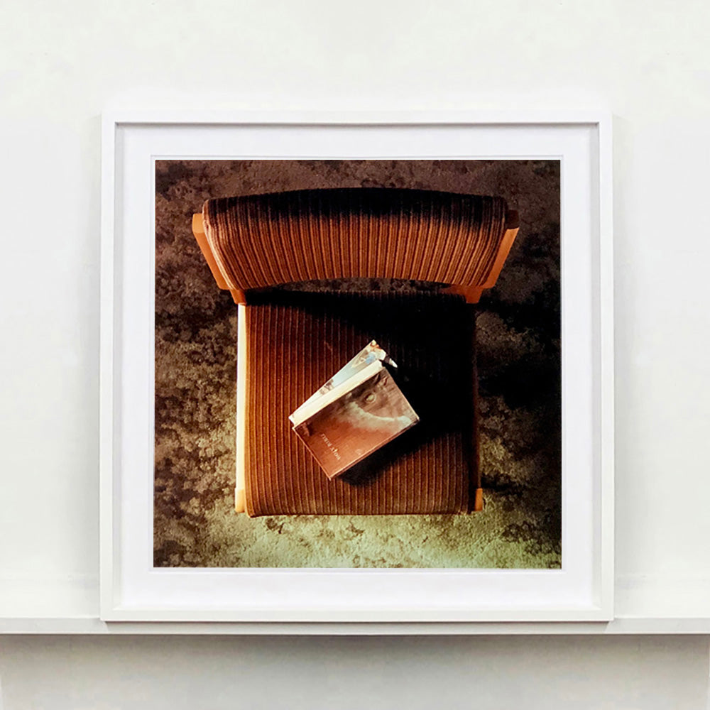 White framed photograph by Richard Heeps. The photograph looks down on a brown padded side chair. On it sits a waterstained and battered brown covered Holy Bible.