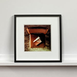 Black framed photograph by Richard Heeps. The photograph looks down on a brown padded side chair. On it sits a waterstained and battered brown covered Holy Bible.