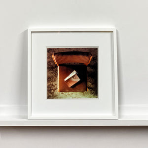 White framed photograph by Richard Heeps. The photograph looks down on a brown padded side chair. On it sits a waterstained and battered brown covered Holy Bible.