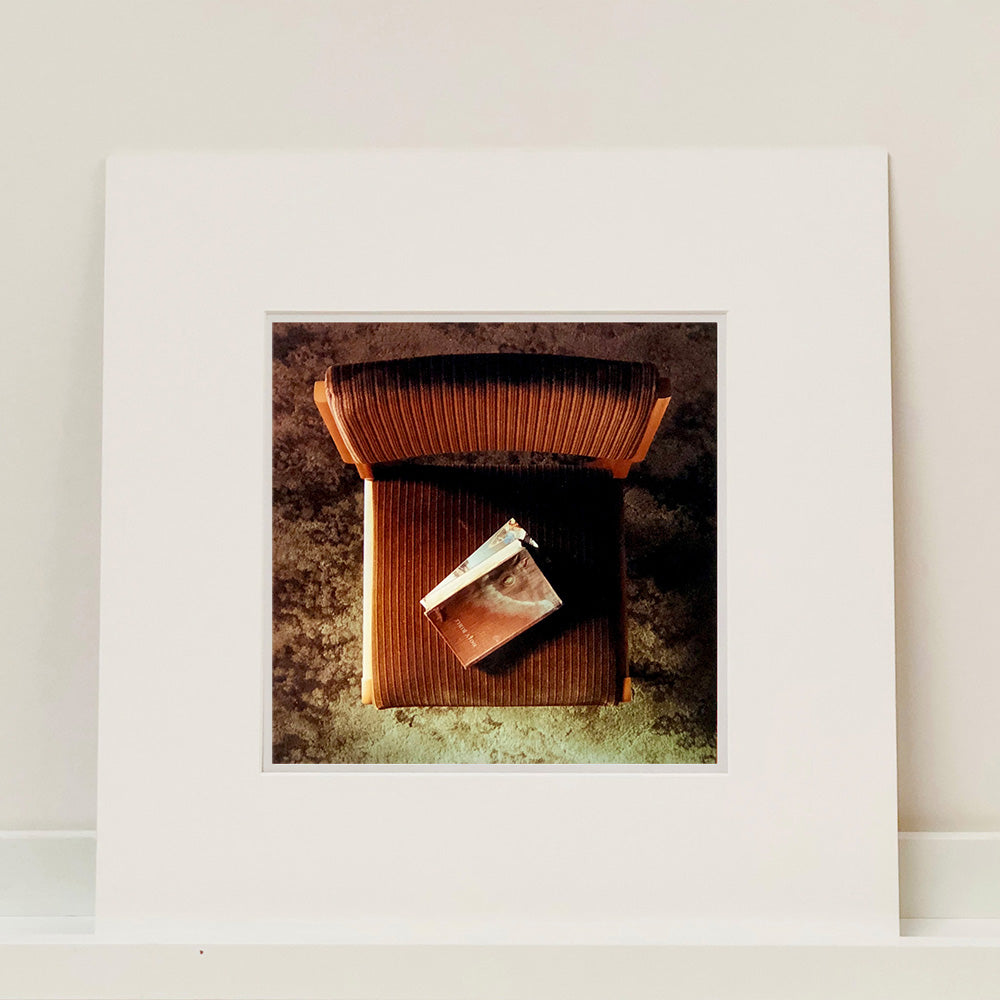 Mounted photograph by Richard Heeps. The photograph looks down on a brown padded side chair. On it sits a waterstained and battered brown covered Holy Bible.