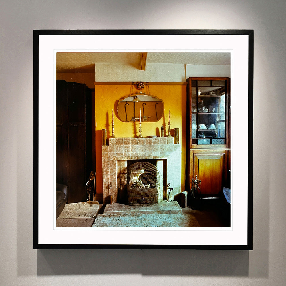Black framed photograph by Richard Heeps. A vintage room with a fireplace with a brown tiled surround sitting in the middle of the photo flooded in daylight. On the mantelpiece sits 3 candlesticks, the walls behind are painted mustard yellow and there is a a decorative mirror. There is a dresser tucked into the recess on the right hand side.