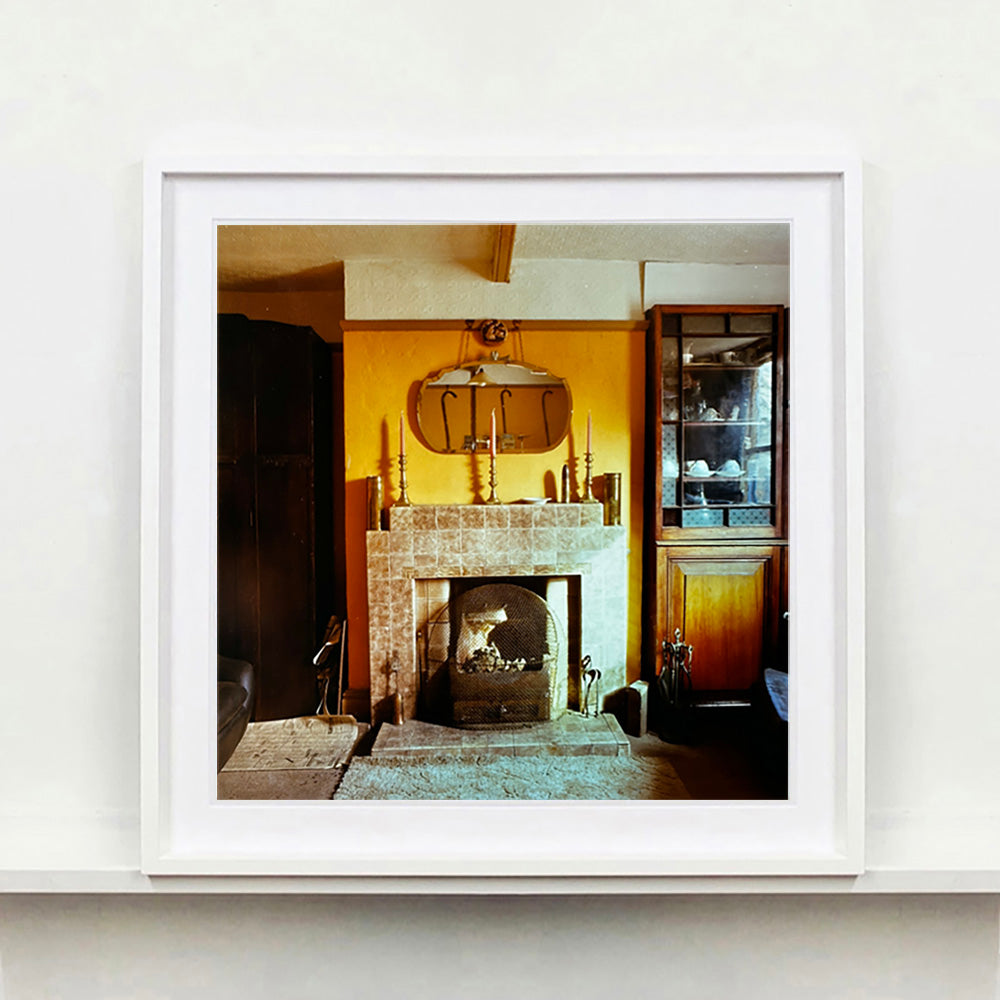 White framed photograph by Richard Heeps. A vintage room with a fireplace with a brown tiled surround sitting in the middle of the photo flooded in daylight. On the mantelpiece sits 3 candlesticks, the walls behind are painted mustard yellow and there is a a decorative mirror. There is a dresser tucked into the recess on the right hand side.