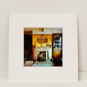 Mounted photograph by Richard Heeps. A vintage room with a fireplace with a brown tiled surround sitting in the middle of the photo flooded in daylight. On the mantelpiece sits 3 candlesticks, the walls behind are painted mustard yellow and there is a a decorative mirror. There is a dresser tucked into the recess on the right hand side.