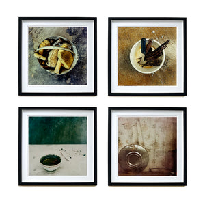Four black framed photographs by Richard Heeps hung in a square. The first photograph is a bucket with sponges, brushes and wooden handled tools sitting in it on a cracked cement floor. The second one is of a selection of wooden handled knives sitting in a white container, the third photograph is a small white rice bowl with a green inside, sitting on a white table with a green wall behind. The fourth photograph is a metal hubcap, sitting against a faded brown flowered curtain.