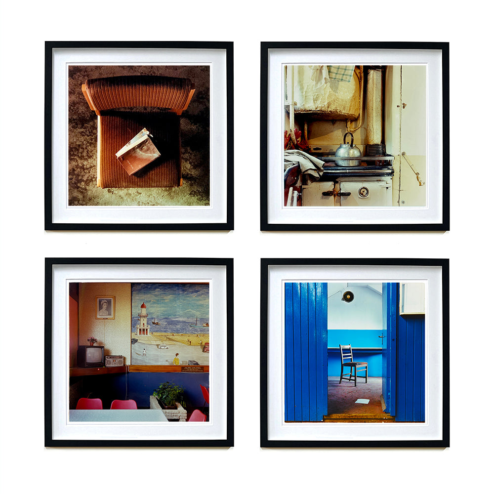 Four black framed photographs by Richard Heeps. The first photograph looks down on a brown padded side chair. On it sits a waterstained and battered brown covered Holy Bible. The second photograph is an old aga with a metal kettle on top. The third is a cafe with a sea side painting on the wall, the fourth is a chair sitting alone in a blue room.