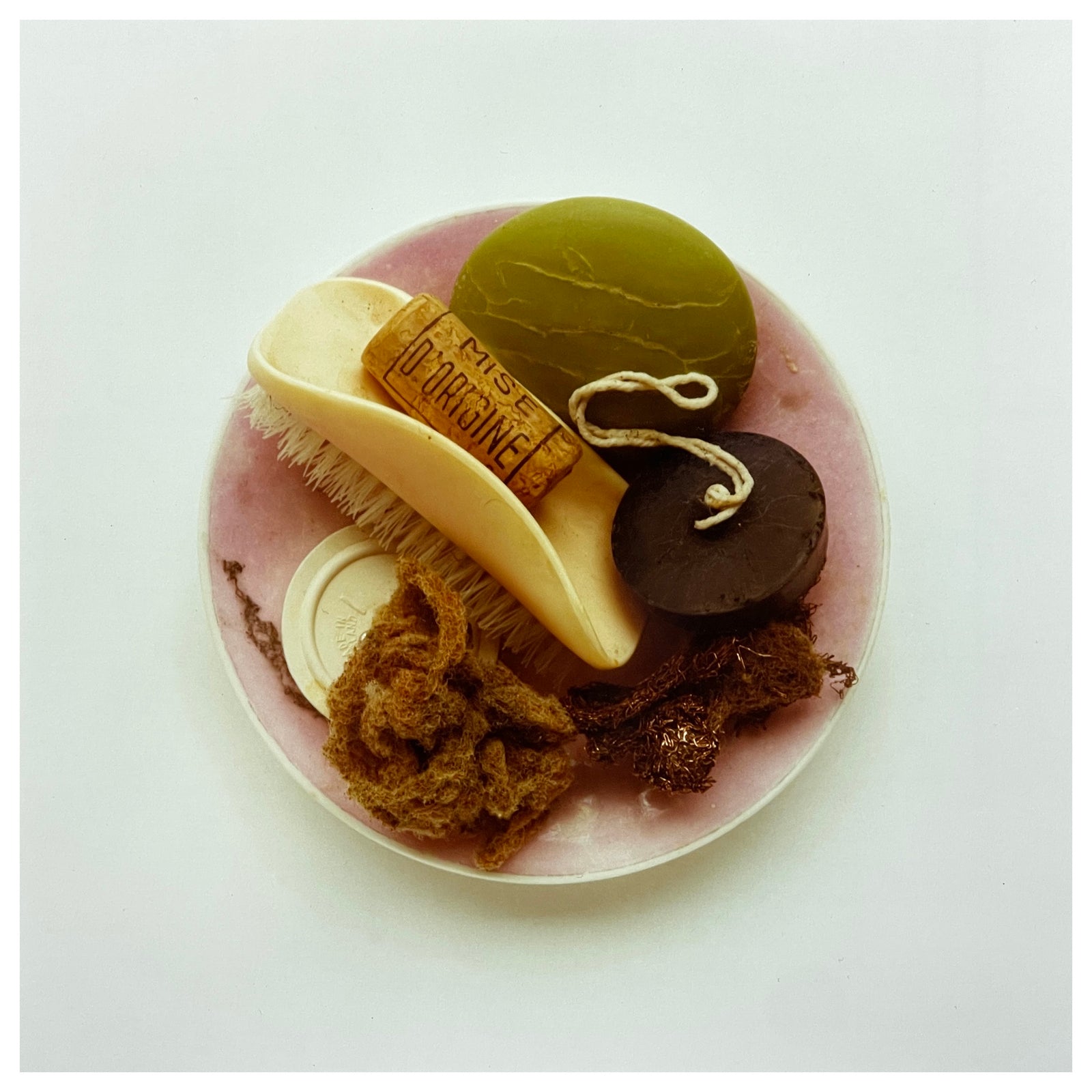 Photograph by Richard Heeps. A vintage pink soap dish sits on a white surface. Looking down onto the soap dish which contains two plugs, green soap, nail brush, cork and a sponge/wire wool combo.