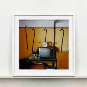 White framed photograph by Richard Heeps. Photograph of a vintage room, a television is on a table with two aerials on top, beside the television are drinks and glasses. Hanging from the picture rail on the yellow wall behind the television are 4 different walking sticks and a bed pan. 