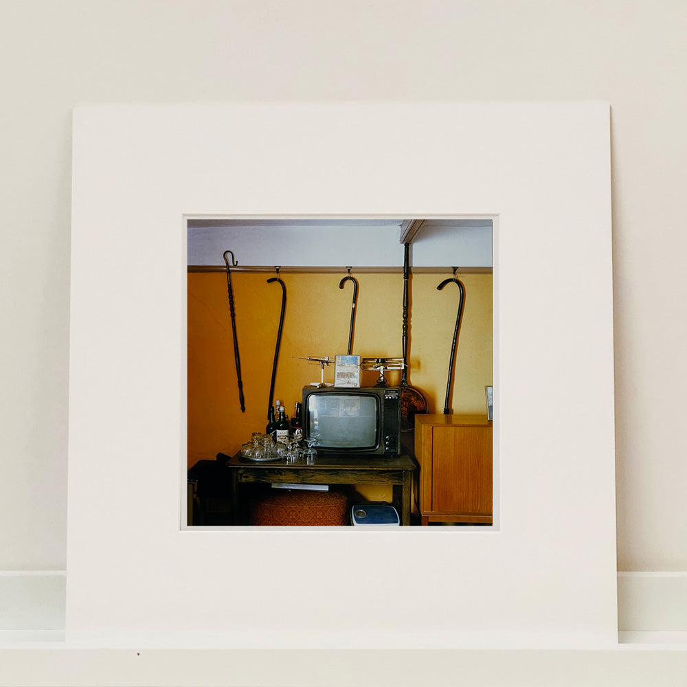 Mounted photograph by Richard Heeps. Photograph of a vintage room, a television is on a table with two aerials on top, beside the television are drinks and glasses. Hanging from the picture rail on the yellow wall behind the television are 4 different walking sticks and a bed pan. 