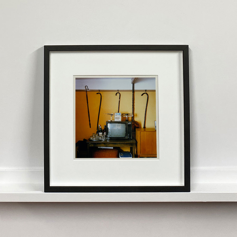 Black framed photograph by Richard Heeps. Photograph of a vintage room, a television is on a table with two aerials on top, beside the television are drinks and glasses. Hanging from the picture rail on the yellow wall behind the television are 4 different walking sticks and a bed pan. 