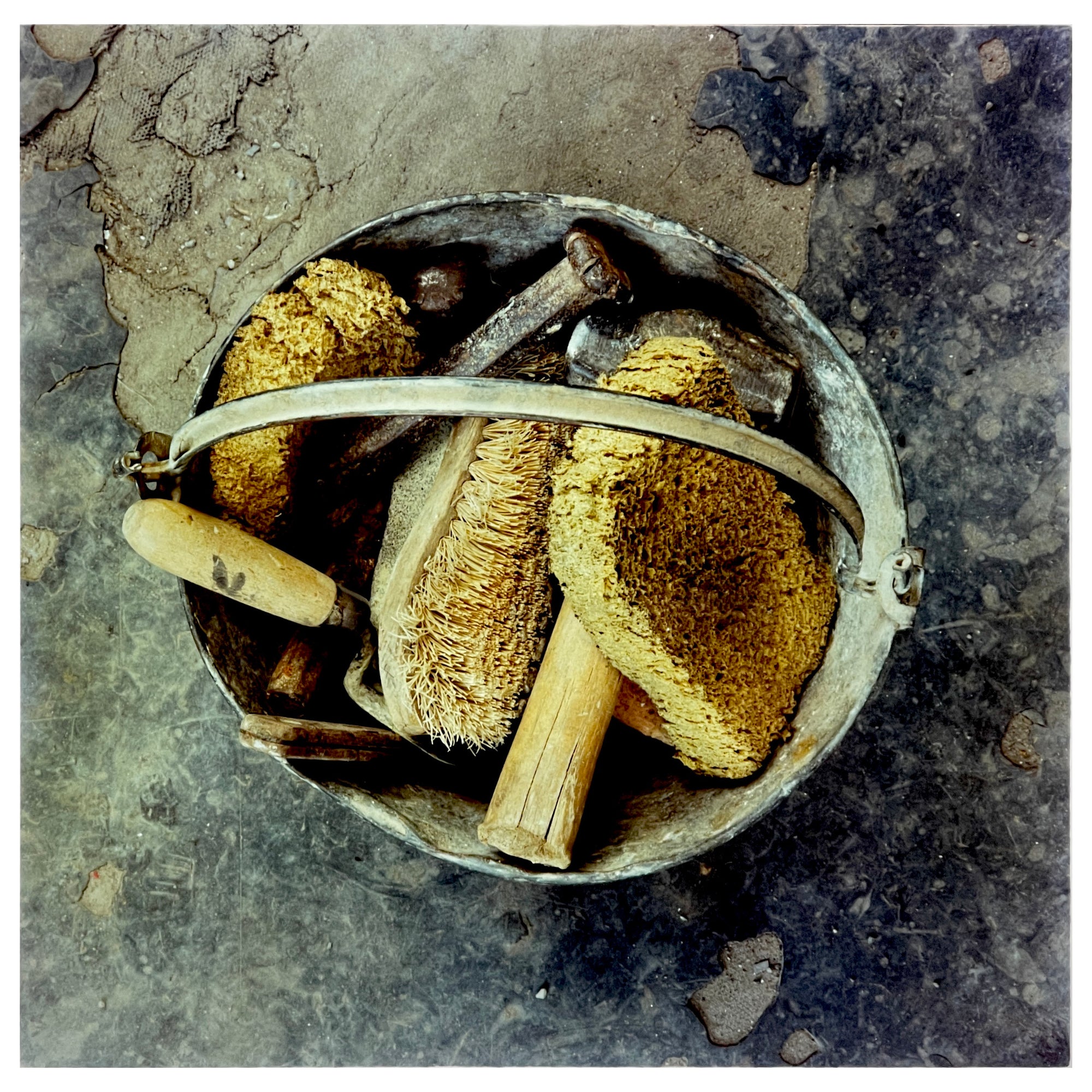 Photograph by Richard Heeps. A bucket with sponges, brushes and wooden handled tools sit in a bucket on a cracked cement floor.