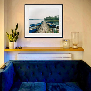A framed Norfolk Broads photograph by Richard Heeps of a wooden dock extending over calm water with blue boats, displayed on a beige wall above a wooden shelf in a modern living room setting.
