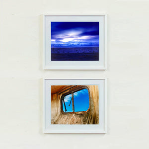 Two white framed photographs by Richard Heeps. The top photograph captures blue light as it hits vast land, mountains and a big sky. The bottom photograph is a blue sky through the window of a dilapidated trailer.