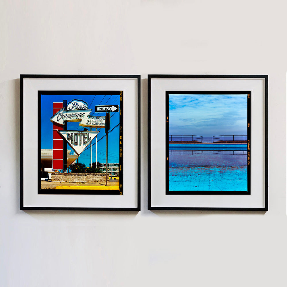 Two black framed photographs by Richard Heeps. On one side the photograph depicts a group of signs on an American Road. Arrow sign pointing to the left to Pink Champagne, pointing down with MOTEL and a one way sign pointing to the right. The Motel signs sit in front of the hotel on a red squared ladder. On the right hand side the photograph is an expanse of water of the most beautiful blues, the scene is cut in half by a blue concrete path, with a wooden and metal fence, with a gap in the middle.