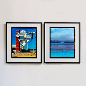 Two black framed photographs by Richard Heeps. On one side the photograph depicts a group of signs on an American Road. Arrow sign pointing to the left to Pink Champagne, pointing down with MOTEL and a one way sign pointing to the right. The Motel signs sit in front of the hotel on a red squared ladder. On the right hand side the photograph is an expanse of water of the most beautiful blues, the scene is cut in half by a blue concrete path, with a wooden and metal fence, with a gap in the middle.