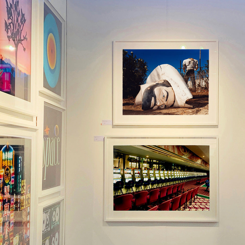 White framed photograph by Richard Heeps. In the foreground of the photograph is a broken off head of a giant fibreglass sculpture of a cowboy (a Muffler Man). In the background set in a deep blue sky is the muffler man's headless torso. The photograph is set amongst some other of Richard Heeps' photographs exhibited at the Affordable Art fair.