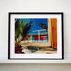 Black framed photograph by Richard Heeps. A colourful but derelict vintage motel sits in the blue Californian sun. Palm trees appear behind and to the side. 