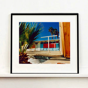 Black framed photograph by Richard Heeps. A colourful but derelict vintage motel sits in the blue Californian sun. Palm trees appear behind and to the side. 