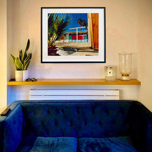 An in situ black framed photograph by Richard Heeps. A colourful but derelict vintage motel sits in the blue Californian sun. Palm trees appear behind and to the side. 