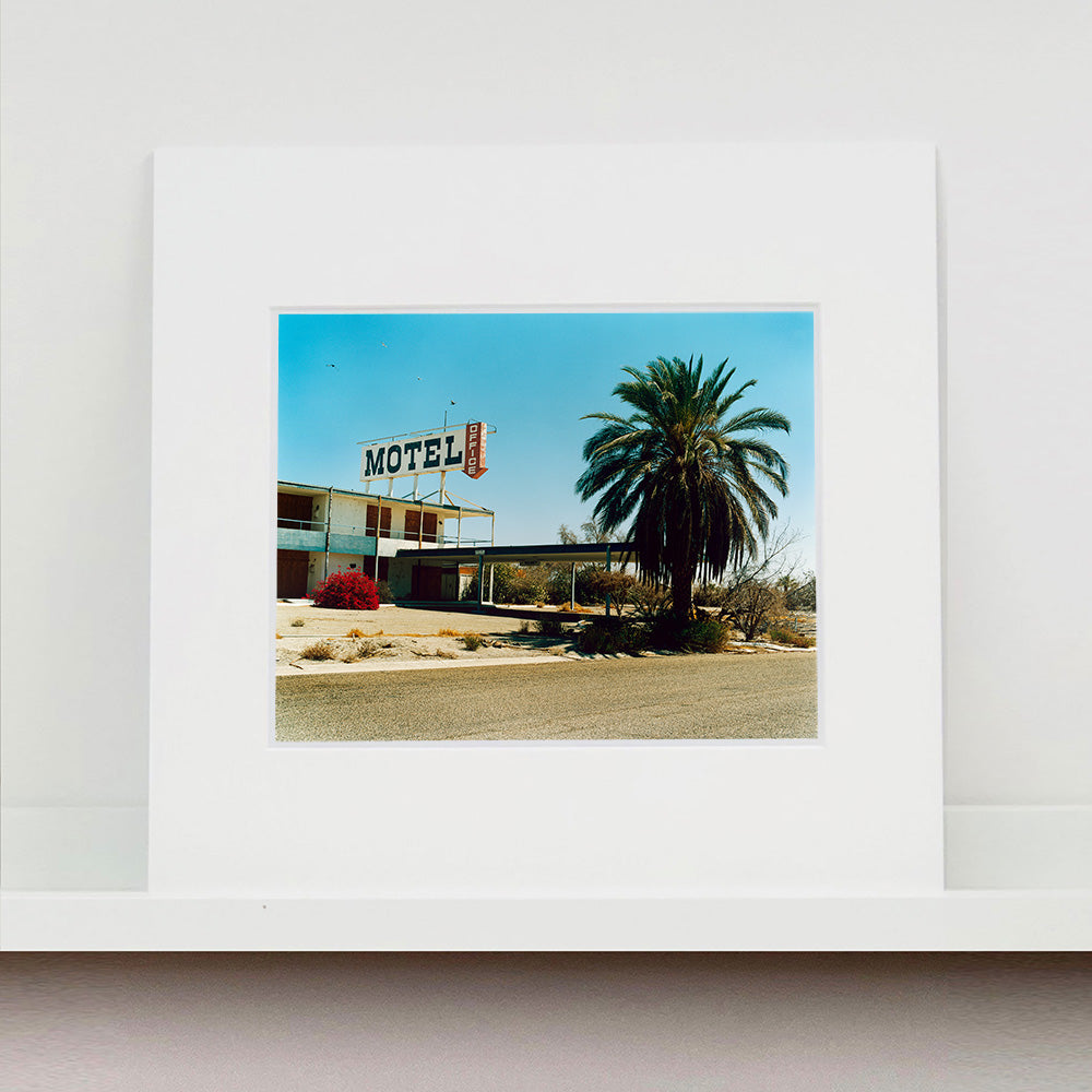 Photograph by Richard Heeps. A derelict motel office sits on a dusty American road. A large palm tree sits at the front of the office's walkway.