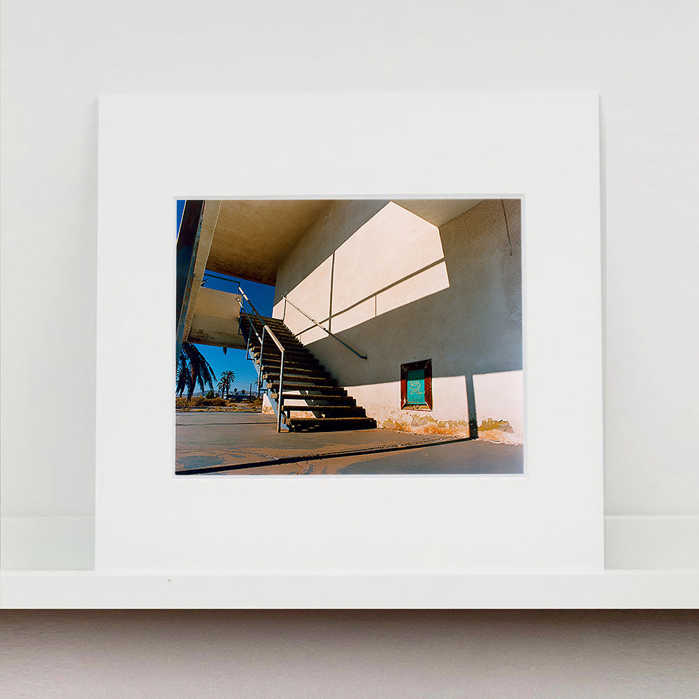 Mounted photograph by Richard Heeps. The photograph has a metal staircase on the outside of a cream colour motel. The staircase has a ceiling but no sides so leads to blue sky. Behind the building is a blue sky and palm trees of the Californian Desert.