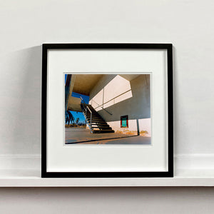 Black framed photograph by Richard Heeps. The photograph has a metal staircase on the outside of a cream colour motel. The staircase has a ceiling but no sides so leads to blue sky. Behind the building is a blue sky and palm trees of the Californian Desert.