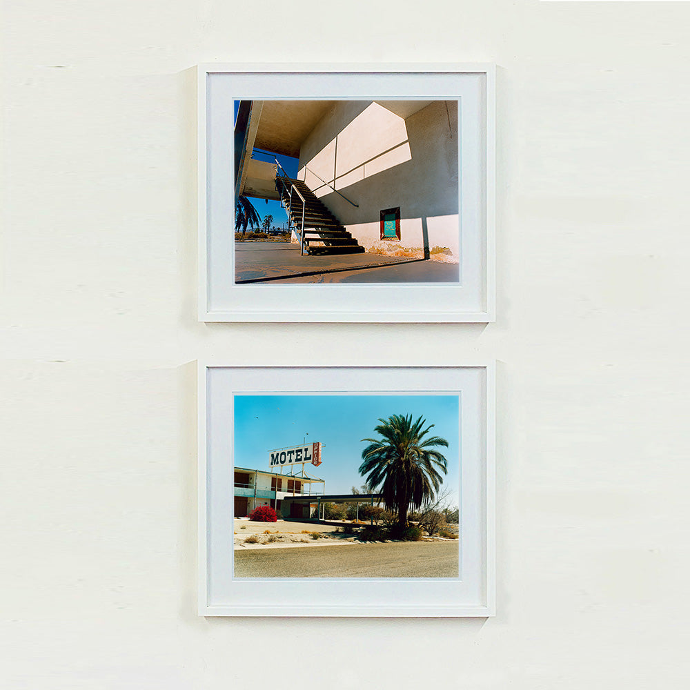 Two white framed photographs by Richard Heeps. The photograph has a metal staircase on the outside of a cream colour motel. The staircase has a ceiling but no sides so leads to blue sky. Behind the building is a blue sky and palm trees of the Californian Desert. The bottom photograph is of a run down motel sitting on a dusty road. At the end of the Motel walkway is a huge palm tree.