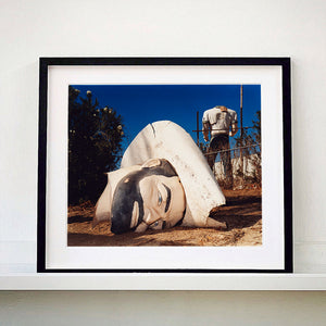 Black framed photograph by Richard Heeps. In the foreground of the photograph is a broken off head of a giant fibreglass sculpture of a cowboy (a Muffler Man). In the background set in a deep blue sky is the muffler man's headless torso.