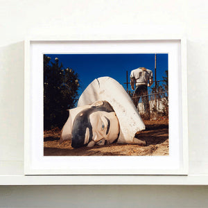 White framed photograph by Richard Heeps. In the foreground of the photograph is a broken off head of a giant fibreglass sculpture of a cowboy (a Muffler Man). In the background set in a deep blue sky is the muffler man's headless torso.