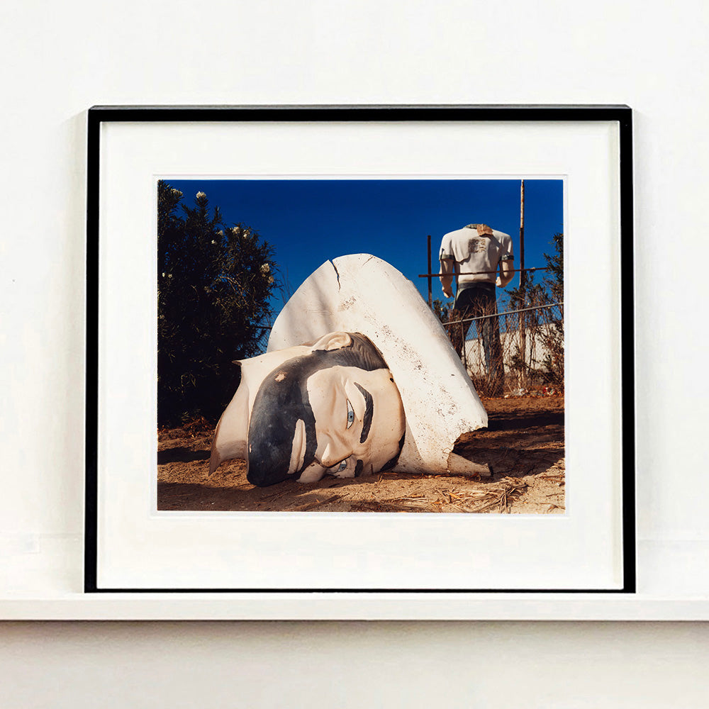 Black framed photograph by Richard Heeps. In the foreground of the photograph is a broken off head of a giant fibreglass sculpture of a cowboy (a Muffler Man). In the background set in a deep blue sky is the muffler man's headless torso.