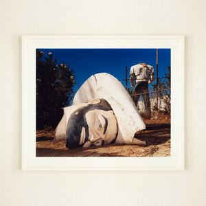 White framed photograph by Richard Heeps. In the foreground of the photograph is a broken off head of a giant fibreglass sculpture of a cowboy (a Muffler Man). In the background set in a deep blue sky is the muffler man's headless torso.