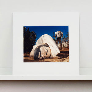 Mounted photograph by Richard Heeps. In the foreground of the photograph is a broken off head of a giant fibreglass sculpture of a cowboy (a Muffler Man). In the background set in a deep blue sky is the muffler man's headless torso.