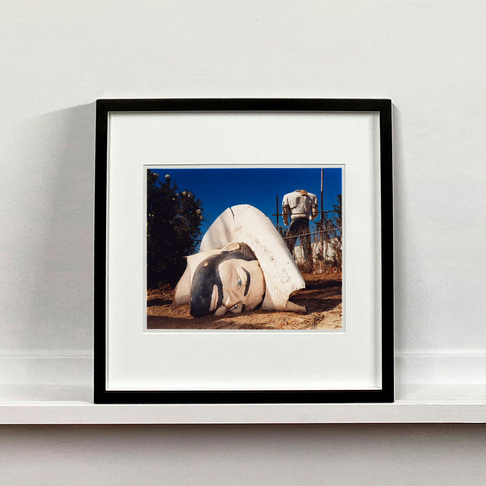 Black framed photograph by Richard Heeps. In the foreground of the photograph is a broken off head of a giant fibreglass sculpture of a cowboy (a Muffler Man). In the background set in a deep blue sky is the muffler man's headless torso.