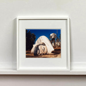 White framed photograph by Richard Heeps. In the foreground of the photograph is a broken off head of a giant fibreglass sculpture of a cowboy (a Muffler Man). In the background set in a deep blue sky is the muffler man's headless torso.