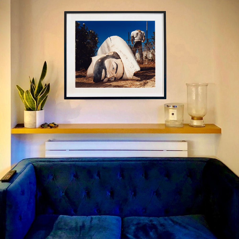 In situ black framed photograph by Richard Heeps. In the foreground of the photograph is a broken off head of a giant fibreglass sculpture of a cowboy (a Muffler Man). In the background set in a deep blue sky is the muffler man's headless torso.